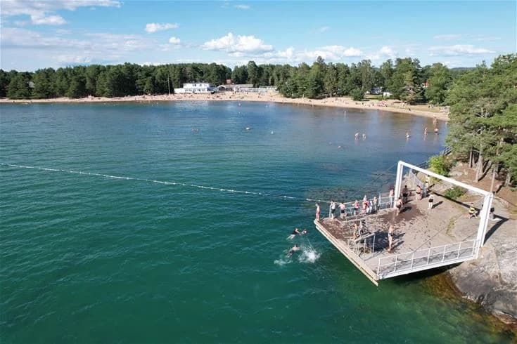 En solig dag på Harge Bad & Camping med utsikt över en sandstrand och det glittrande havet i bakgrunden. Besökare njuter av det vackra kustområdet och det finns människor vid stranden som kopplar av och deltar i olika fritidsaktiviteter.