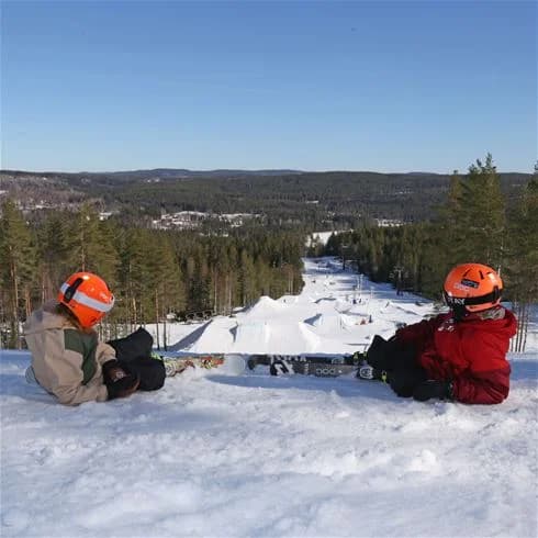 En snötäckt skidbacke i Bjursås Berg & Sjö med människor som åker skidor. Winterlandskap med barrträd i bakgrunden under en klar himmel.
