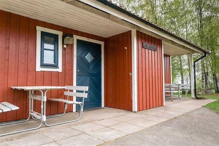 En trästuga med en bred dubbeldörr, omgiven av träplankor och en enkel trästol under skuggorna. Byggnaden påminner om en rustik campingmiljö, typisk för Mösseberg camping.