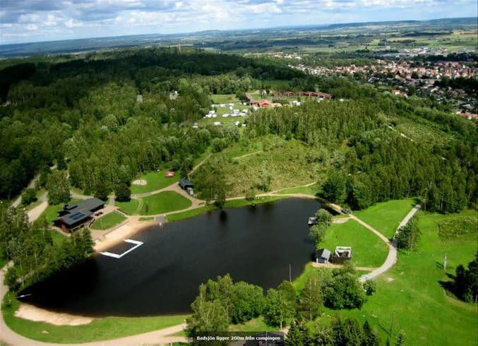 Bild av Mösseberg Camping med ett naturskönt område som visar träd, växter och en damm. Gräsmattor och ängar sträcker sig intill vattnet, vilket skapar en fridfull parkmiljö.