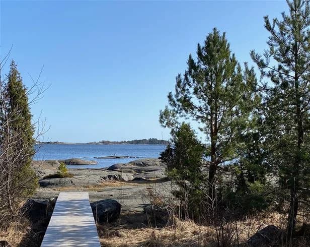 Vy över Ängskärs havscamping med en blå himmel som speglas i det närliggande vattnet. Kusten pryds av grönskande växtlighet och stenar, vilket skapar en harmonisk och naturskön miljö.