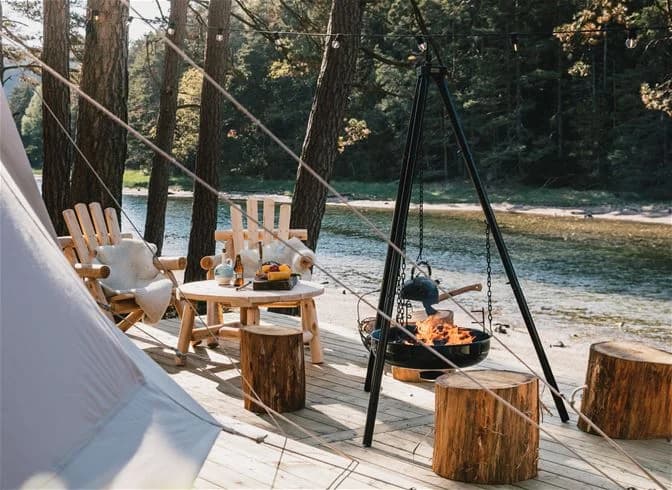 En campingplats med tält och stugor omgiven av tät skog. I förgrunden syns en lägereld omgiven av några stockar att sitta på. Scenen ger en känsla av friluftsliv och avslappnad naturupplevelse.