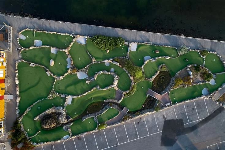 Flygbild över Lagunen Camping & Stugor som visar en vackert anlagd minigolfbana omgiven av grönska och artificiellt gräs, med blommande trädgårdsytor och landskapsdesign i en parkmiljö.