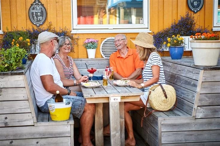 En träveranda hos Lagunen Camping & Stugor med utemöbler och blomkrukor. Ett matbord är dekorerat med växter och serverad mat, omgiven av träfärgade stolar och omgivande grönska.