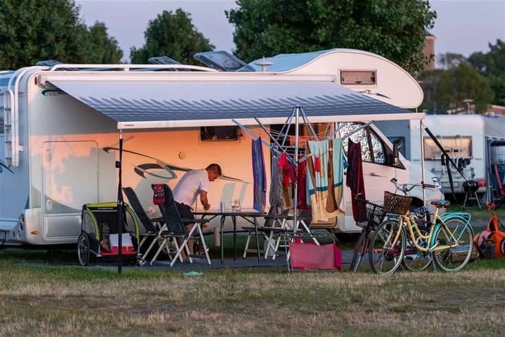 En campingplats med husbilar och tält under solskydd, människor som njuter av fritidsaktiviteter i skuggan av en markis, och stolar uppställda för avkoppling.
