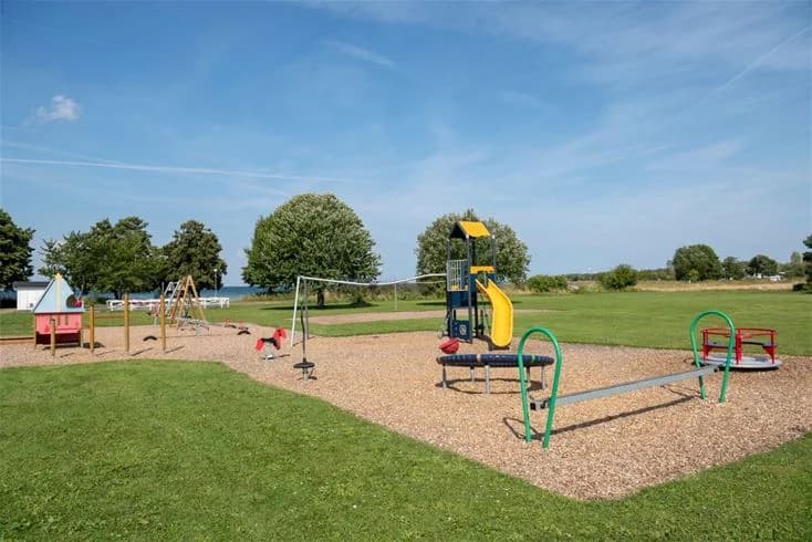 En solig dag vid Gröndals camping & stugor med en blå himmel och grönskande gräsmatta. Lekplatsutrustning syns på området, omgiven av öppna ytor och skugga från träden.