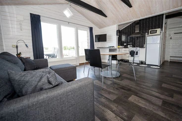 Inuti en trästuga med träväggar och trägolv. Rummet är möblerat med en dagbädd och en stol. Det finns en dörr som leder ut från stugan.