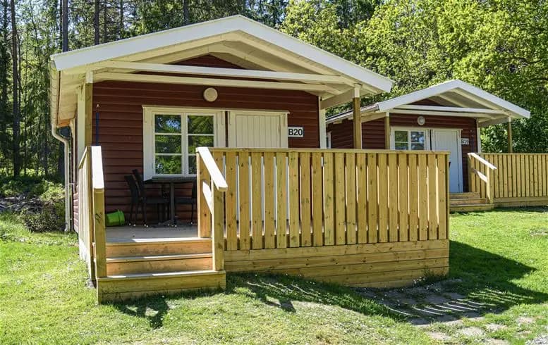 En trästuga med träpaneler belägen i en lummig trädgård vid Ursand Resort & Camping, omgiven av gröna växter och skuggiga områden.