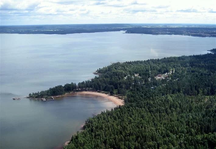 Bilden visar en naturskön kuststräcka vid Ursand Resort & Camping med ett klart vattendrag i förgrunden, omgiven av lummiga träd och en sandstrand längs kusten.