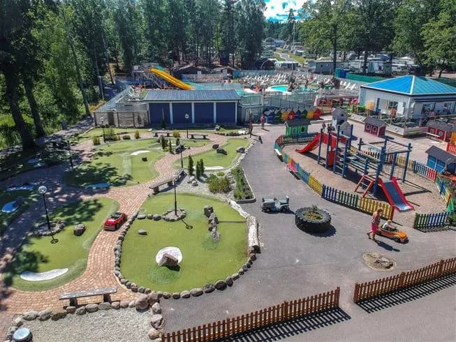 En solig dag vid Ursand Resort & Camping där familjer njuter av fritid och lek i en lummig park med lekutrustning.