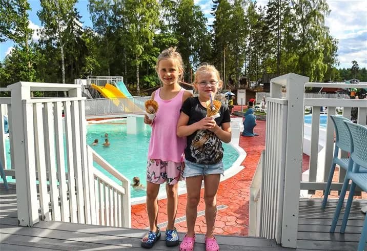 En grupp människor njuter av solen vid en stor utomhuspool på Ursand Resort & Camping. I förgrunden syns flera barn i badkläder som leker och har roligt i vattnet. Bakgrunden visar en livlig semesteratmosfär med solstolar och parasoller vid en välbesökt campinganläggning.