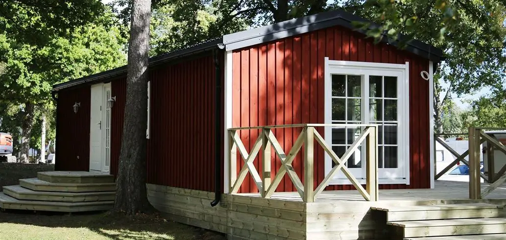 En trästuga med mörkt trä, som står på campingen Ursand Resort. Stugan har en enkel design med en träfällningsdörr och omges av grönska, vilket ger en rustik och naturnära känsla.