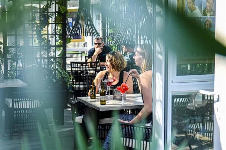 En utomhusservering med bord och stolar under skuggande parasoll på Ursand Resort & Camping. I bakgrunden syns grönska och fler sittgrupper.