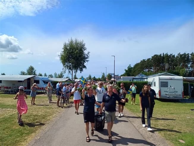 Människor njuter av en solig dag på Båstad camping, omgivna av husbilar och grönska. En livlig stämning med besökare som promenerar och deltar i utomhusaktiviteter.