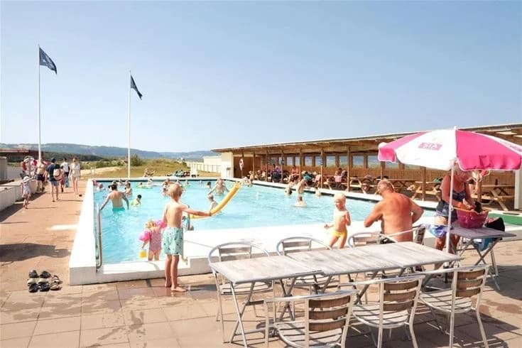 Bild av Båstad Camping som visar en solig dag vid en pool. Familjer njuter av semester, omgivna av utemöbler och gröna områden. En avslappnad atmosfär vid en populär semesterort.