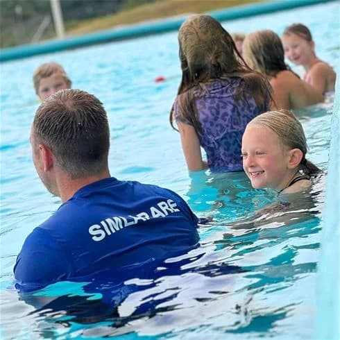 En familj njuter av en sommardag vid en pool på Båstad camping, barn leker och simmar medan solen skiner, vilket skapar en avkopplande semestermiljö.
