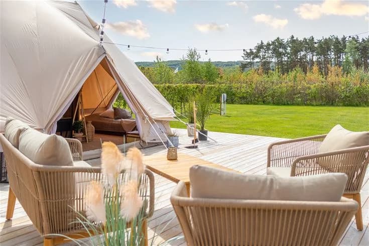 Bild av en campingplats i Båstad med utomhusmöbler och tält. Det finns stolar och ett bord under ett parasoll och ett canopy-tält syns i bakgrunden.