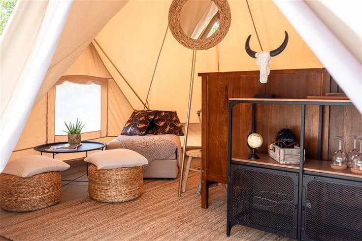 En campingplats med ett tält uppsatt, inrett med bekväma möbler som stolar, kuddar och en korg. Tältet har ett varmt och inbjudande utseende med träfläckar och textilier. En krukväxt kompletterar den hemtrevliga atmosfären.