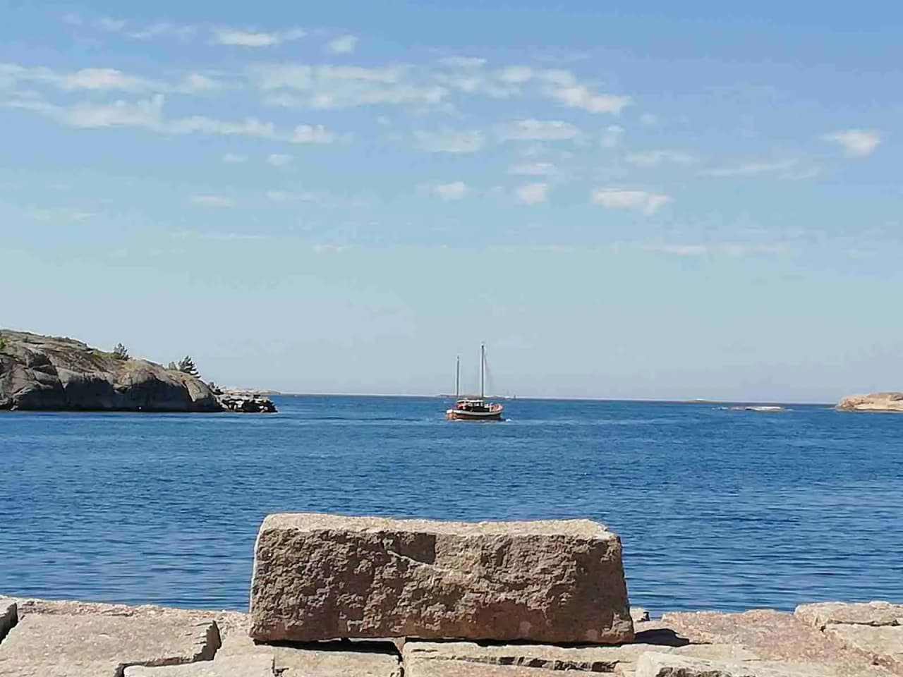 Utsikt över Hunnebostrands camping och stugor med klarblått vatten och kustlinje i bakgrunden. Klippor och horisont syns tydligt, medan en båt färdas på det lugna havet.