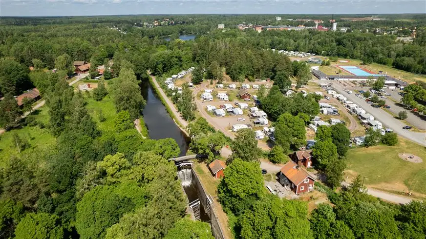 Flygbild över Skantzö Bad & Camping som visar en grönskande park med träd, en kanal, och campingområden. Parkeringsplats och intilliggande semesterort syns även i bilden.