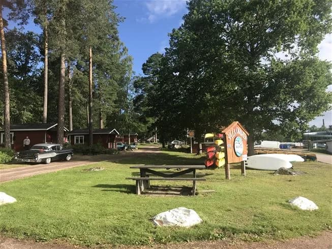 En grönskande campingplats med stugor omgivna av träd och välklippta gräsmattor. Det finns en skuggig trädgård och en smal infart som leder in mot området.