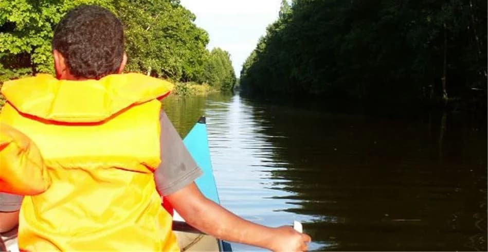En grupp människor åker på en motorbåt på en sjö eller flod, alla bär flytvästar. Bakgrunden visar frodig grönska kring vattnet, vilket tyder på en naturskön miljö vid Skantzö Bad & Camping.