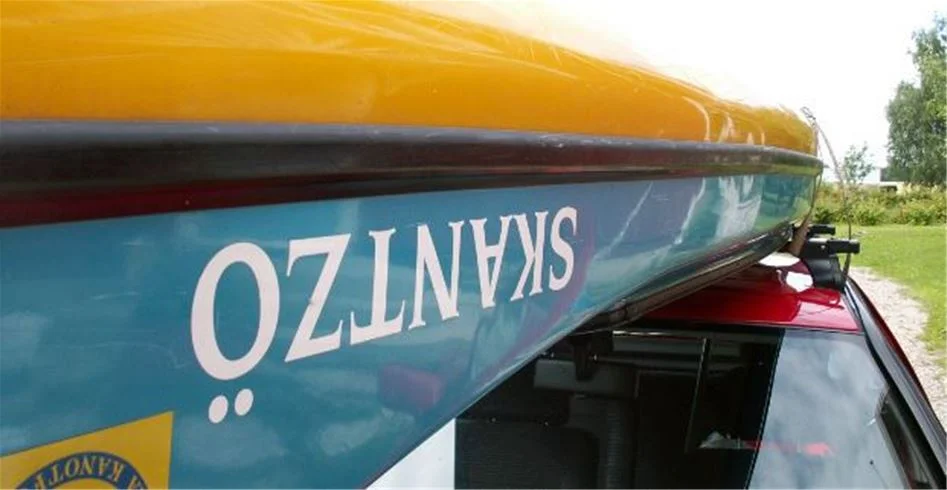 En utsikt över Skantzö Bad & Camping med flera uppställda kajaker på ett biltak bredvid ett staket. Området erbjuder vattenaktiviteter och naturskön omgivning.