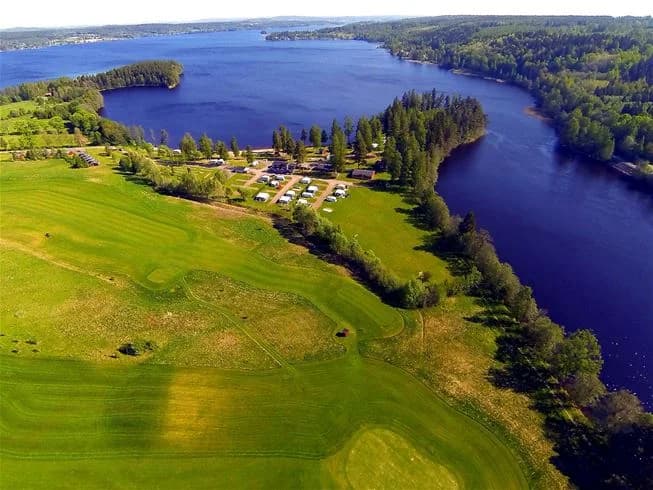 Flygbild av Degernäs Camping vid en sjö med klart vatten och omgivande grönska. Reflektioner av himlen syns på vattenytan, vilket skapar en lugn och naturskön atmosfär.