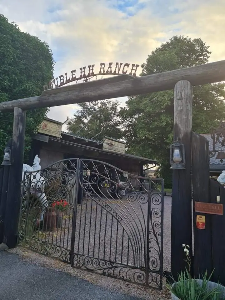 En smidesgrind markerar ingången till en lummig och välskött trädgård med staket som omger platsen, sannolikt en ställplats vid namn Wild West. I bakgrunden syns trädgårdslandskap och utomhusstrukturer som skapar en inbjudande atmosfär.