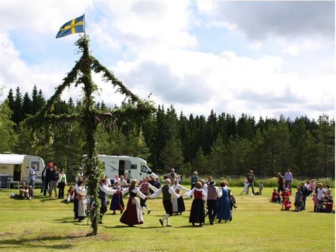 En grupp människor samlade runt en midsommarstång på Gröna Backe Camping & Stugor under en traditionell svensk midsommarfirande. Färgglada band pryder stången och omgivningen visar en festlig och gemenskaplig atmosfär.
