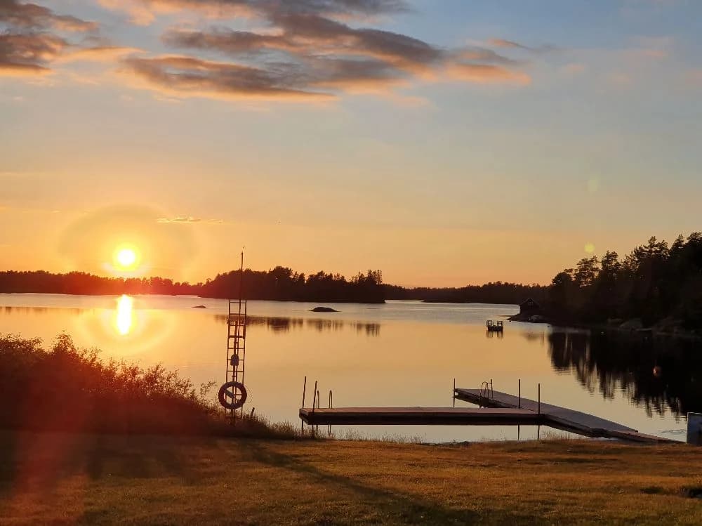 En bild av Tyrislöt camping med en vacker solnedgång över vattnet. Himlen är färgad i dämpade pastelltoner och horisonten syns i fjärran, medan strandkanten avtecknar sig mot det lugna vattnet.