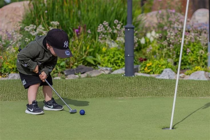 En golfspelare svingar sin klubba på en grön golfbana vid Grebbestadfjorden. Golfutrustning syns på gräsmattan och det är en solig dag med klarblå himmel.