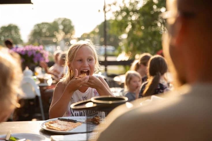 En grupp glada människor äter och umgås vid ett bord på Kneippbyn Resort Visby, med färgglada bordstillbehör och mat omkring dem.