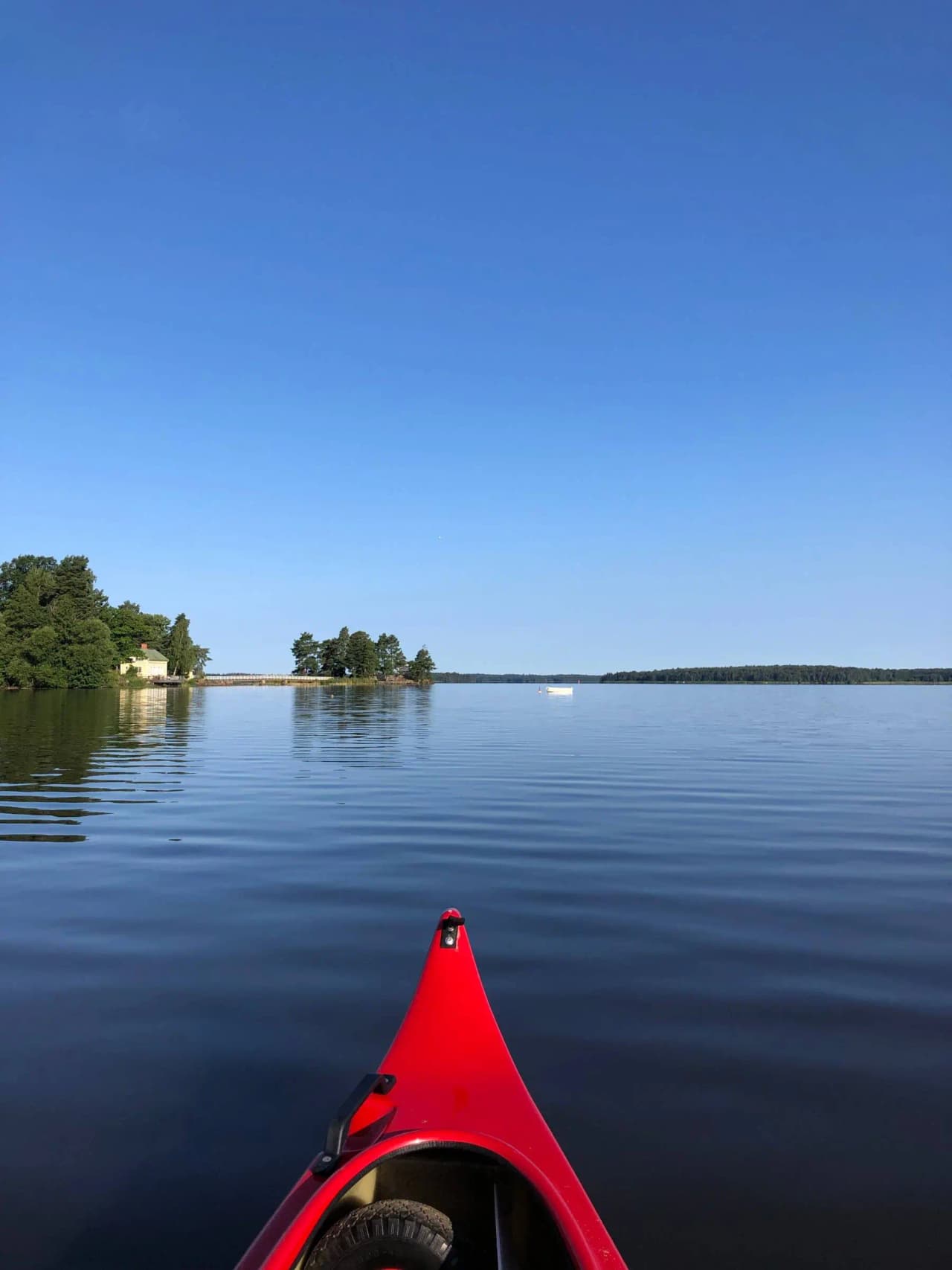 En stilla sjö vid Mälarbadens camping där en person paddlar en kajak på det glittrande vattnet. Flera andra småbåtar syns i bakgrunden, omgivna av grönskande natur.