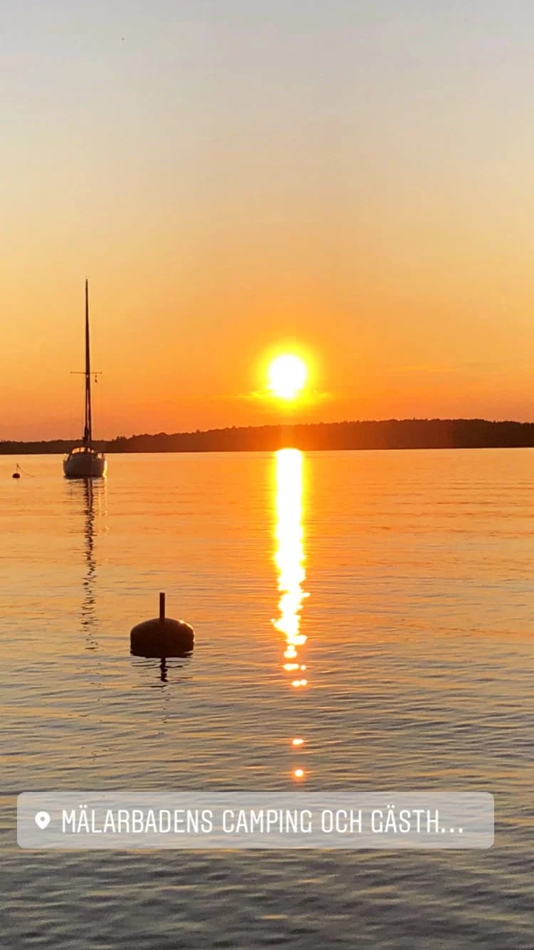 Solnedgång vid Mälarbadens camping, med en båt på vattnet och en orange himmel vid horisonten.