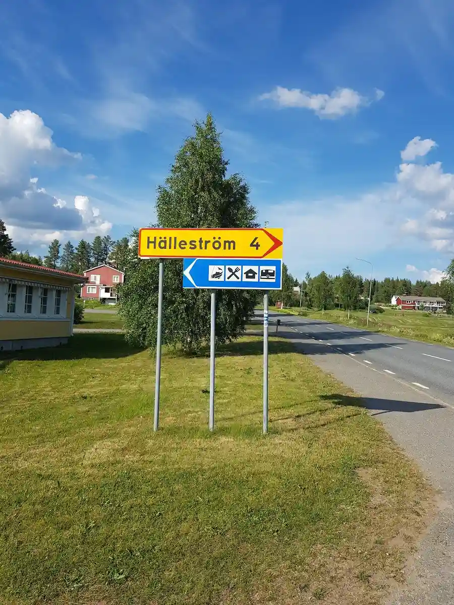 En grön gräsmatta vid Fällfors camping med en vägskylt på en stolpe bredvid en asfalterad väg kantad av frodig gräsyta.