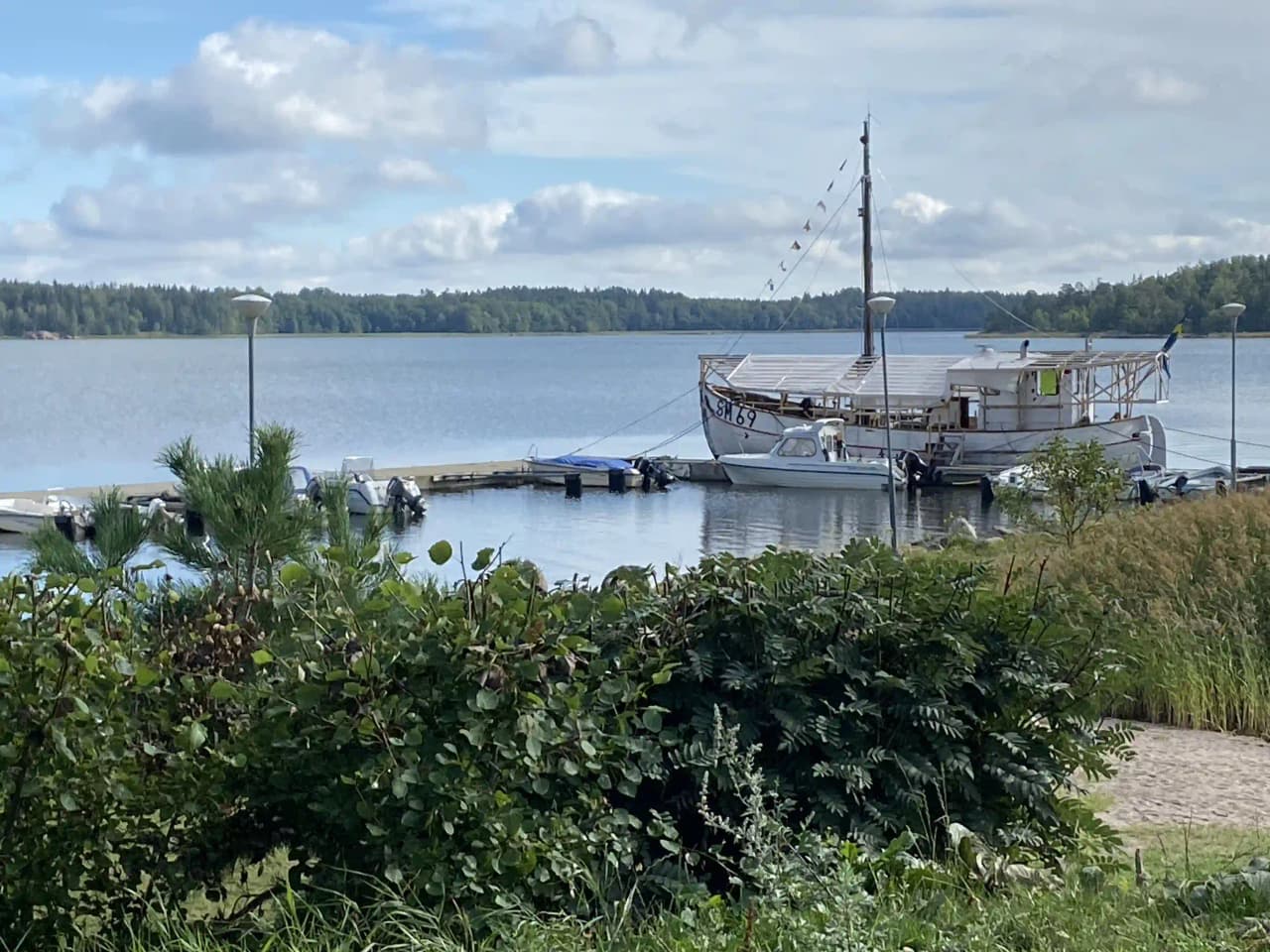 En sommardag vid Hargshamns camping med båtar förtöjda vid en glittrande sjö, omgivna av moln och grönska i ett kustnära landskap.