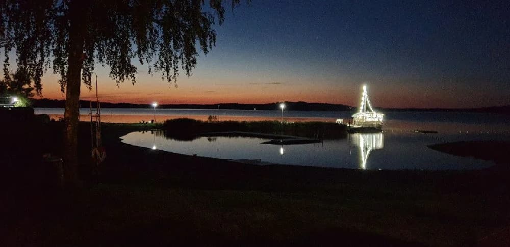 En stilla sjö i skymningen med speglingar på vattnet, omgiven av mörk siluett av träd och berg vid Hargshamns camping.