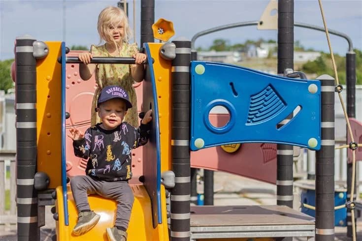 Barn som leker vid en lekplats på Grebbestadfjorden, med rutschkanor och annan utelekutrustning. De bär färgglada barn- och småbarnskläder och verkar ha roligt i den friluftslika miljön.