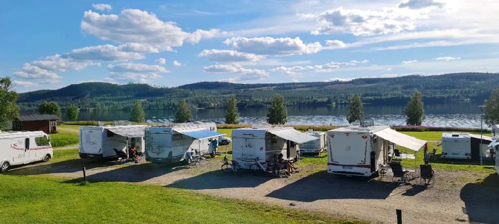En husvagn står parkerad på en grön äng under en molnig himmel, nära en lugn sjö omgiven av skog. Perfekt för camping och avkoppling i naturen.