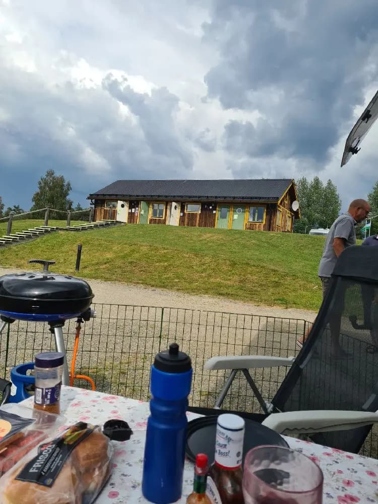 En campingplats med tält och en presenning som ger skugga över ett picknickbord och utemöbler på en gräsmatta. I bakgrunden syns ett lantligt hus. Perfekt för en avkopplande vistelse i naturen.