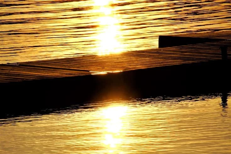 En solnedgång som reflekteras i en stilla sjö vid Vallsnäs camping, vilket skapar en mjuk orange och gul färgpalett över vattnet.