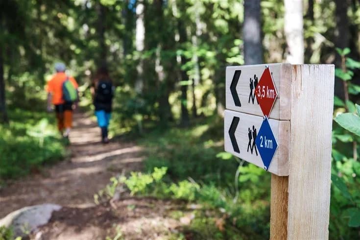 Bild av en skogsstig i Hultsfred Strandcamping, omgiven av tät skog och trädstammar. En skylt syns bredvid stigen, vilket tyder på närhet till en naturreservat eller rekreationsområde. Atmosfären är fridfull och naturnära.