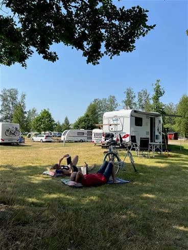 En campingplats med tält och husbilar uppställda på grönt gräs. Träd omger området, och människor njuter av fritiden i soligt väder på Hultsfred strandcamping.