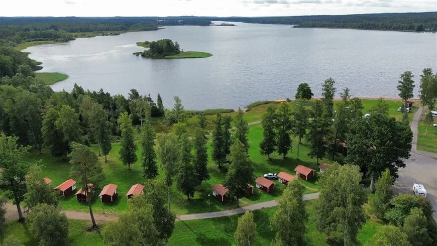 En naturskön vy över Hultsfred Strandcamping med en glittrande sjö omgiven av frodiga växter och grönska. I bakgrunden syns några stugor längs vattenkanten i ett lantligt område.
