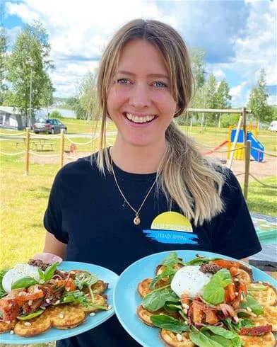 En grupp människor äter frukost vid ett campingbord utomhus, omgivna av gröna träd och en klar himmel. På bordet finns tallrikar med olika frukter och enklare rätter. Stämningen är avslappnad och gemytlig.