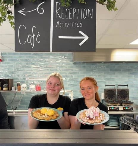 En grupp människor sitter vid ett matbord på Hultsfred strandcamping, omgivna av frukost- och snabbmat. Det är en avslappnad atmosfär med fokus på gemenskap och matglädje.
