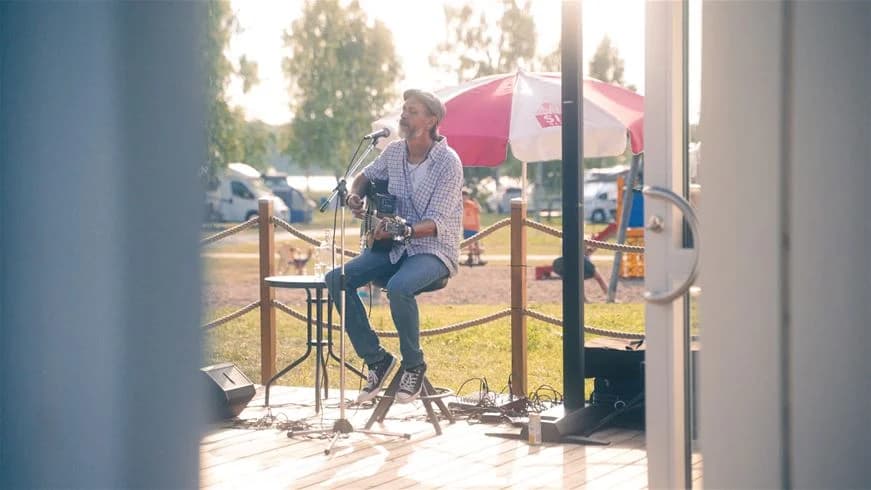 En person sjunger i en mikrofon på en scen vid Hultsfred strandcamping, omgiven av musikutrustning och underhållningstillbehör.