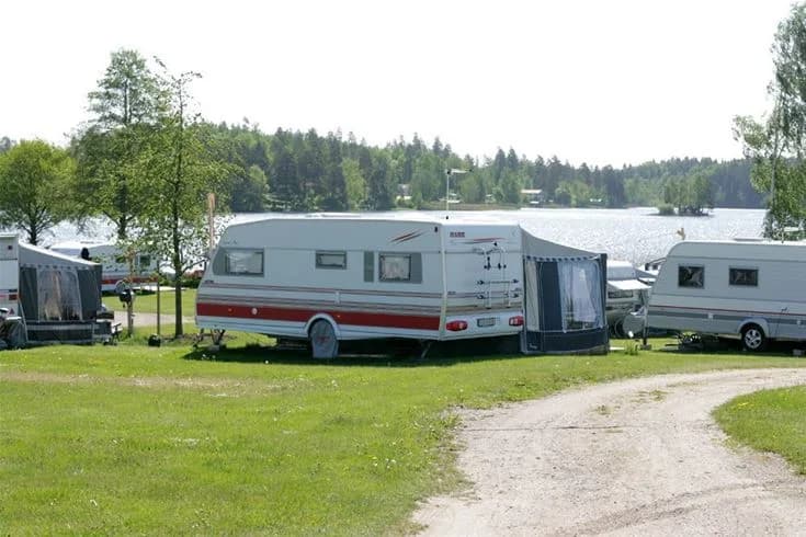 En mobil hemvagn och en campingbil står parkerade på gräset i Djulö camping, omgivna av gröna träd och en rymlig park.