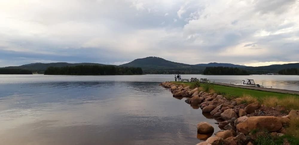 En naturskön campingplats vid vatten med utsikt över en horisont där himlen möter ett bergigt landskap. Vattenväg och moln syns i bakgrunden.
